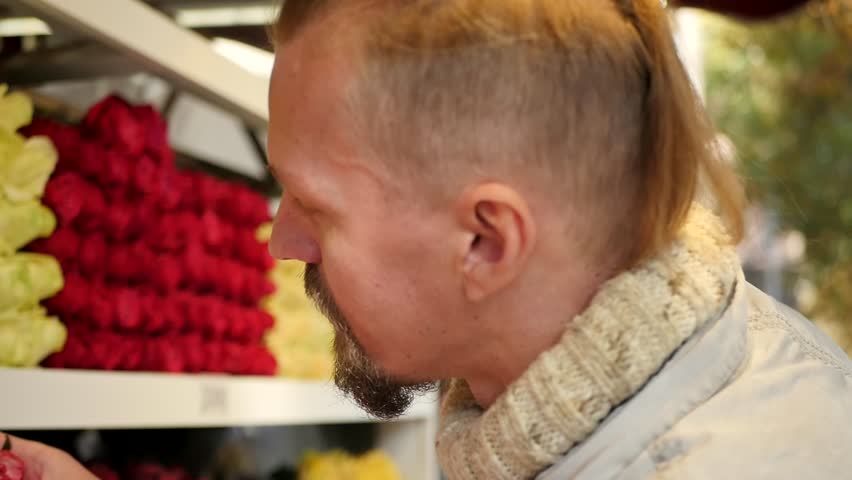 A young handsome bearded man with a fashionable hairstyle buys one rose in a flower shop and, after smelling the flower, gives it to the camera, holding it in his hand. Valentine