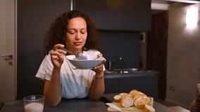 Slow motion. Multi ethnic curly haired young adult brunette, charming pensive woman taking her breakfast in the morning, eating cereals with milk, dreamily looking away, standing by kitchen counter - Powered by Shutterstock - Get 15% off with code: PIKWIZARD15