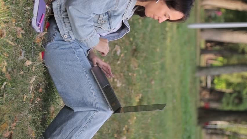 Young woman with a laptop sitting on the grass in the park on an autumn day. Vertical video