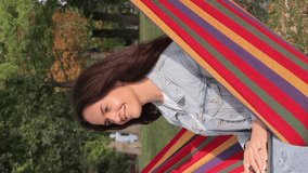 Beautiful woman with a laptop in the garden sitting on a hammock, green background. Vertical video