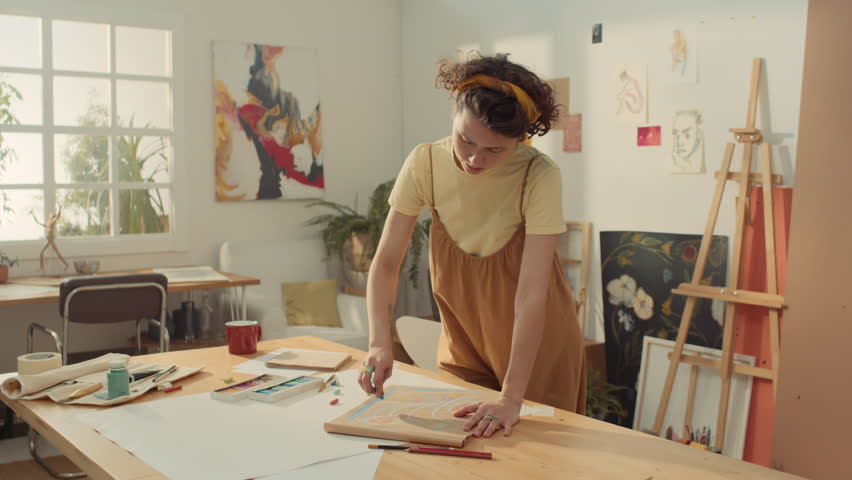 Medium slowmo portrait shot of cheerful young Caucasian woman in jumpsuit, with curly hair drawing sketch with pastel in sketchbook while working in studio, then looking up at camera and smiling Royalty-Free Stock Footage #3403097503