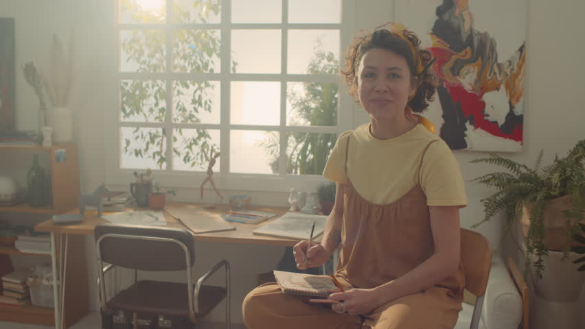 Medium overexposed portrait shot of cheerful young Caucasian female artist with curly hair, in jumpsuit sitting on high chair in workshop, holding sketchbook and pencil, and smiling for camera Royalty-Free Stock Footage #3403109433