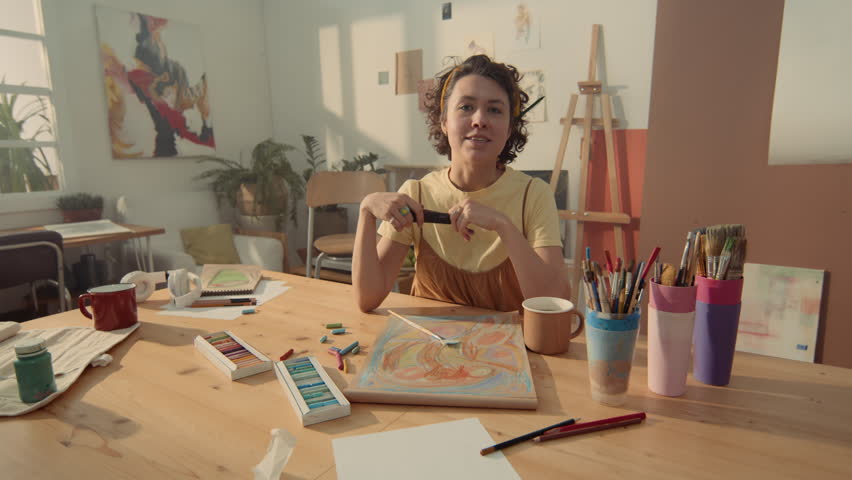 Medium shot of young biracial female art teacher sitting at table in workshop, with sketchbook, pastel, brushes and paints, and beginning painting lesson while filming social media content Royalty-Free Stock Footage #3403109745