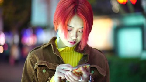 Modern teenage Chinese girl uses smart phone. Non traditional rebellious young Asian woman with colored hair uses phone on street at night. Alternative punk youth downtown in evening.
