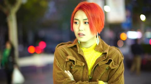 Modern teenage Chinese girl pick up and uses smart phone. Non traditional rebellious young Asian woman with colored hair uses phone on street at night. Alternative punk youth downtown in evening.