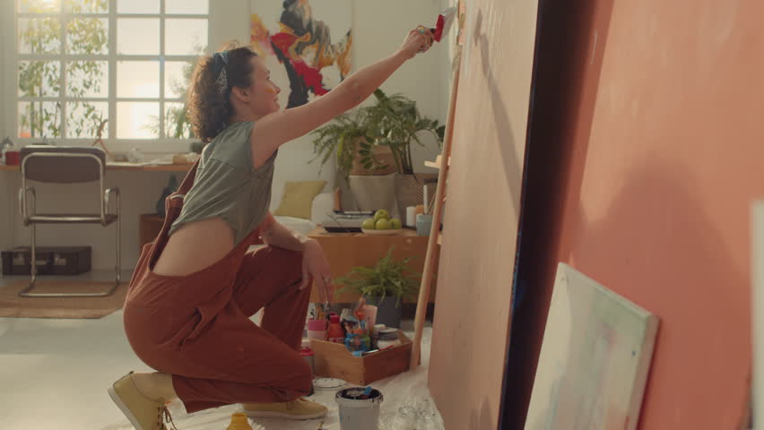 Full side shot of enthusiastic Caucasian female painter in jumpsuit squatting on floor in workshop and drawing abstract lines on plywood sheet with wide brush Royalty-Free Stock Footage #3403130725