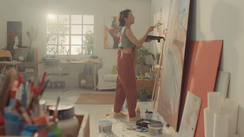 Full side shot of female decorator in jumpsuit standing in studio, holding tray with paint and drawing abstract lines on plywood sheet with wide brush Royalty-Free Stock Footage #3403131493