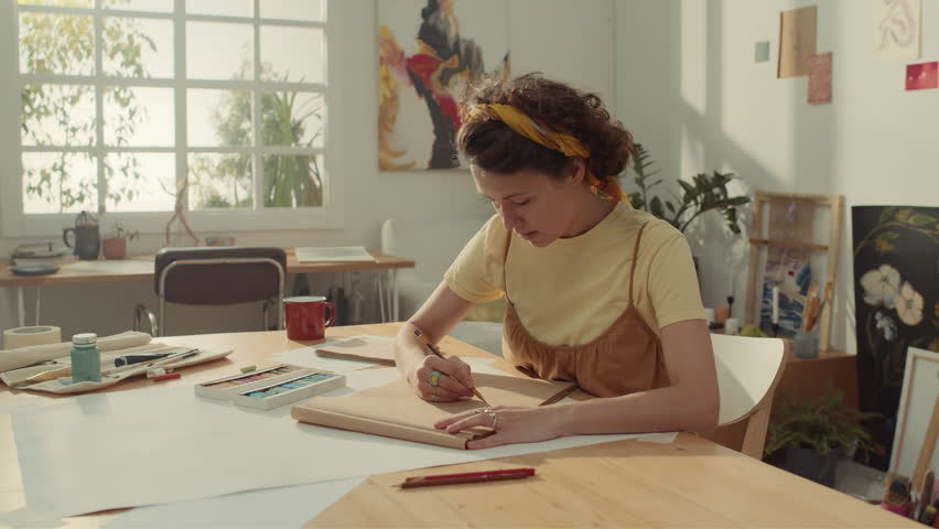 Medium shot of young Caucasian woman with dark curly hair sitting at table in artists workshop and drawing something with pencil in brown sketchbook for future creative masterpiece Royalty-Free Stock Footage #3403134775
