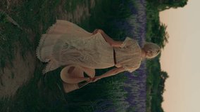 A young blond woman in a white lace dress, and straw bag walks in the beautiful field
