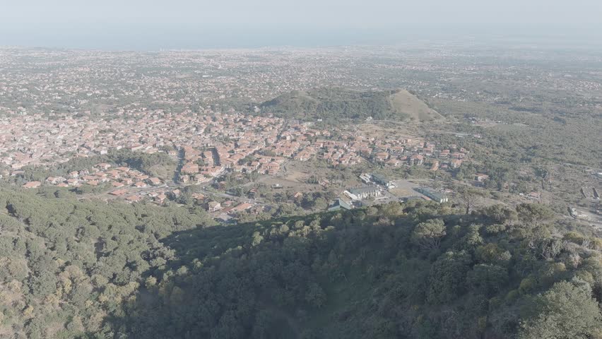 Nicolosi, Sicily, Italy. Twin volcanic cones covered in dense pine forest rise above a sprawling Mediterranean town, captured from an elevated aerial perspective. The two conical craters, known locally as Monti Rossi, were formed during the 1669 eruption of Mount Etna and are now blanketed in evergreen woodland that contrasts with the reddish-brown rooftops of the settlement below. The camera glides smoothly over the forested slopes, revealing the vast urban grid of Nicolosi stretching toward the hazy coastline in the distance. Hazy daylight softens the horizon, creating a calm and expansive atmosphere typical of a warm summer morning. The scene illustrates the dramatic coexistence of volcanic geology and human habitation on the southern flank of one of Europe