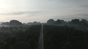 A video from a drone slowly flying from a long straight road with cars moving back and forth in the morning, gradually ascending until it reveals a distant view of forests, mountains, and mist in the 