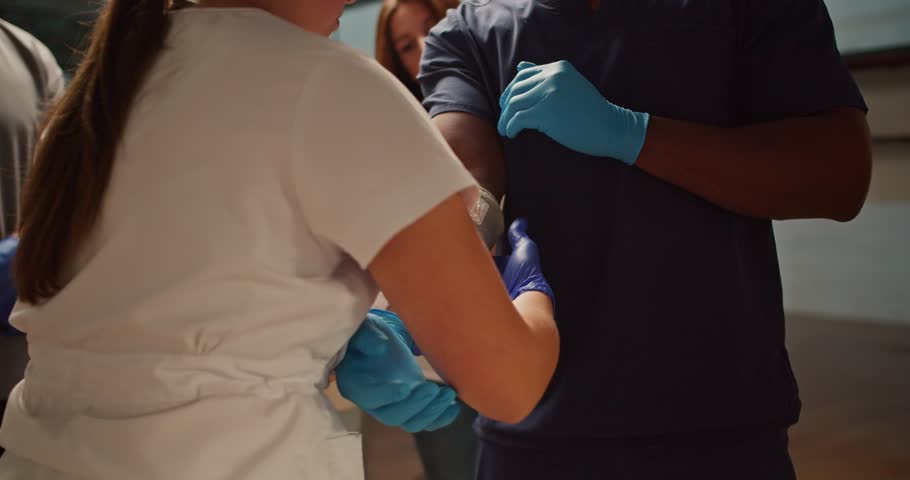 Close-up shooting: A professional female nurse in a white medical uniform puts a bandage on the arm of her assistant - a Black man in a blue medical uniform. Practical training for first aid training Royalty-Free Stock Footage #3403335573