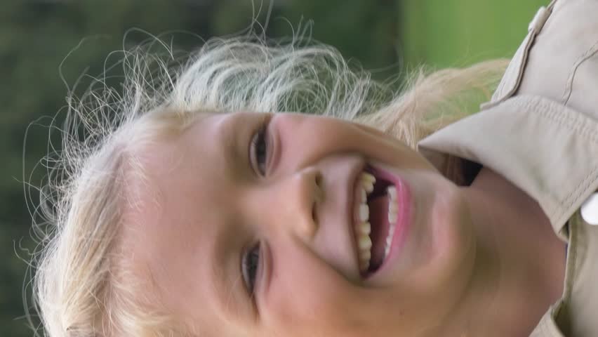Little preschooler caucasian girl with blonde hair in jacket is laughing and grinning in green park. Slow motion. Vertical Video