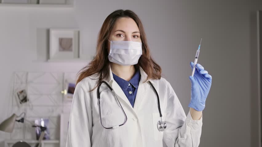 A female doctor in a mask and gloves looks into the camera and holds a syringe filled with liquid. Medicine and Pharmaceuticals Royalty-Free Stock Footage #3403476395