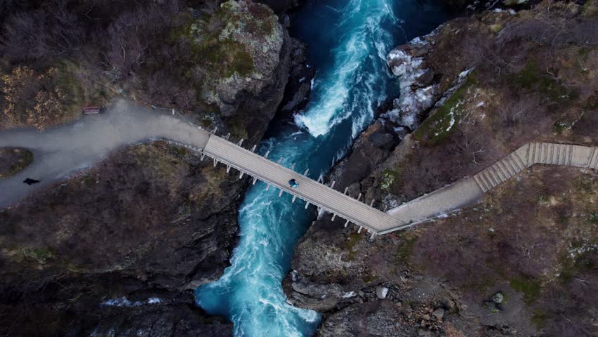Couple standing on impressive bridge over wild river and waterfall in volcanic landscape, Aerial top down rotate