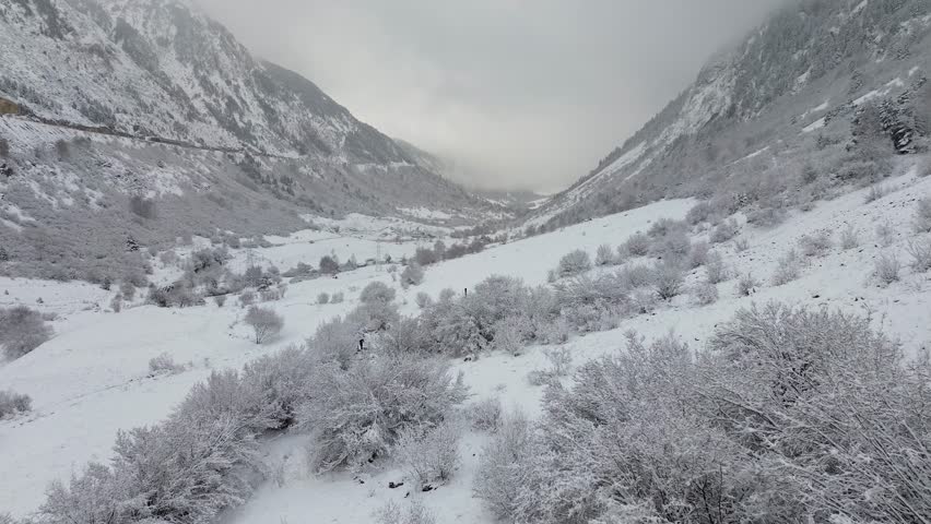 Drone shot of a snow-filled valley in the mountainous Pyrenees of Spain with a small rustic house at the end on a snowy day.