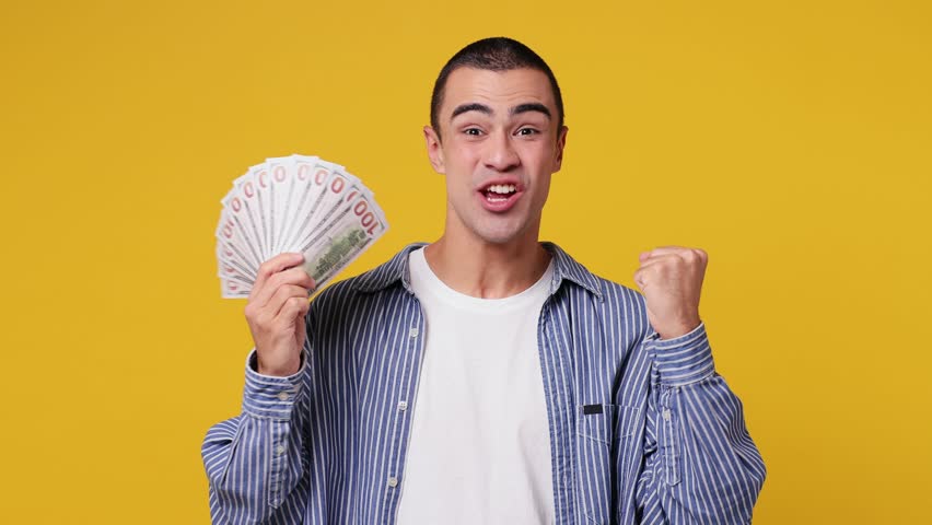 Happy young middle eastern man he wear blue shirt white t-shirt hold in hand fan of cash money in dollar banknotes doing winner gesture celebrate isolated on plain yellow background studio portrait