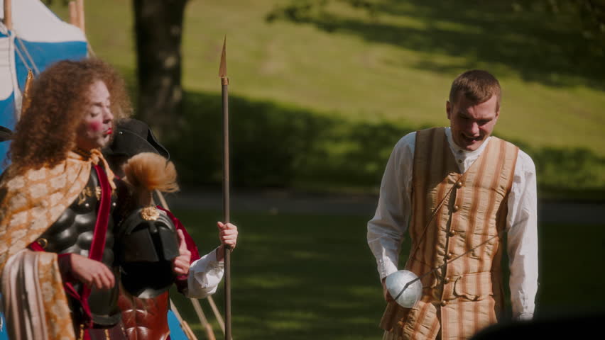 Amidst a medieval setting, an actor dressed in period clothing holds a rapier, engaging in conversation with another actor portraying a medieval general