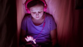 Child girl in the evening under a blanket with headphones flipping through a tablet playing games. High quality 4k footage