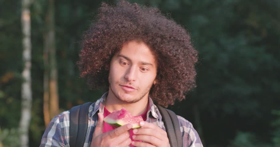 This clip features a young North African man with curly hair, delighting in the simple pleasure of eating a juicy slice of watermelon. His expressions of enjoyment, captured against a backdrop of lush Royalty-Free Stock Footage #3403704155
