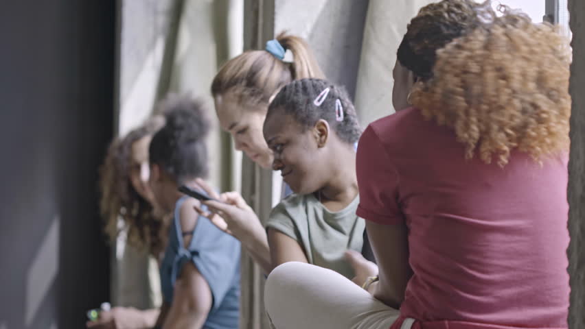 PAN of multiethnic group of beautiful young women sitting on windowsill and chatting while resting after fitness class