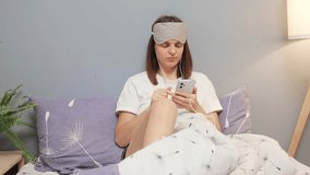 Cheerful woman wearing pajama and sleeping mask using mobile phone for online communicating in social networks enjoying funny video in internet browsing web pages