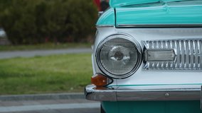 Front round one headlight and false grille of vintage white turquoise car in parking lot on cloudy day. Unrecognizable man in black clothes is walking along path in background. Details of old car. - Powered by Shutterstock - Get 15% off with code: PIKWIZARD15