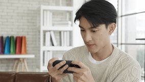 Happy asian man gamer competition play video game online with smartphone in living room at modern house. Esport streaming game online, holds his smartphone with both hands while gaming.