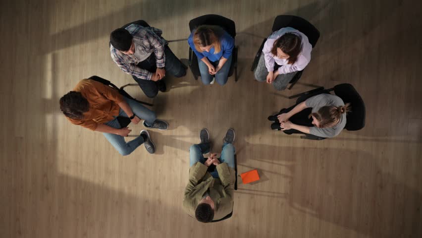 In the shot above, a group of people and their mentor are sitting on a chair. They have come for therapy. They are sitting in a circle writing something in a notebook with a disabled person Royalty-Free Stock Footage #3404063883