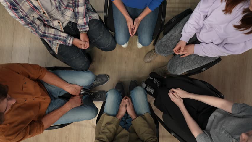 In the frame above sits on a chair in the center, middle aged man, psychologist, mentor. Opposite sits his group they have a meeting, therapy. They are holding hands and talking, communicating Royalty-Free Stock Footage #3404065579
