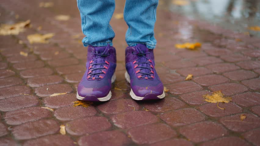 closeup female legs in sport shoes, sneakers. woman walking at rainy autumn day in park. Rain drops Falling On puddle with yellow maple leaves reflection on city street sidewalk.