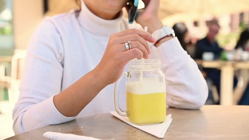 Close-up woman talking on smart phone amd mixing healthy organic juice using a drinking straw, enjoying her weekend outdoor, relaxing in a cozy cafe against unrecognizable blurrred people background