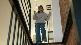 A beautiful girl in headphones descends the stairs in her modern house. As she goes down, she dances and is infinitely joyful in her good mood. High quality 4k footage
