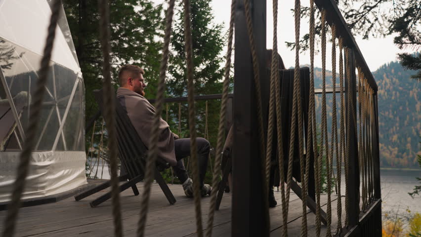 Young couple sits on terrace at eco-hotel. Woman and man enjoy accommodation with opportunity to observe fall nature. Wife sits with husband in armchairs at twilight
