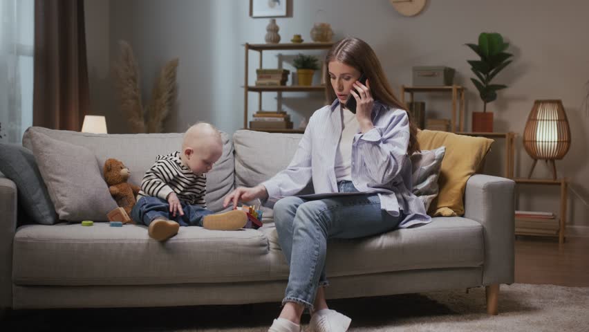 Woman talking by phone and looking after child. Mother looking at work paper. Female resolving work issues with kiddo. Woman managing business while on maternity leave. Kiddo playing with mom. Royalty-Free Stock Footage #3404201969