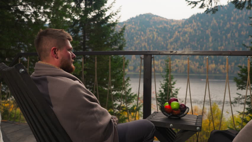 Man enjoys nature view from glamping terrace. Bearded guy wrapped with plaid sits in comfortable armchairs by round tent house. Relaxation in nature