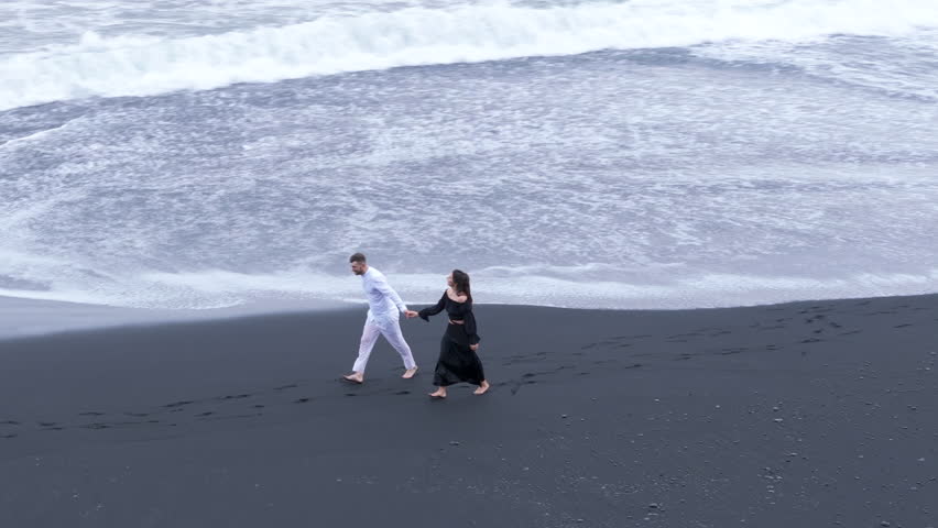 Man in white suit and lady in black long dress running happily on Reynisfjara shore, foaming waves of sea washing their bare feet. Beautiful contrast. High quality 4k footage Royalty-Free Stock Footage #3404345253