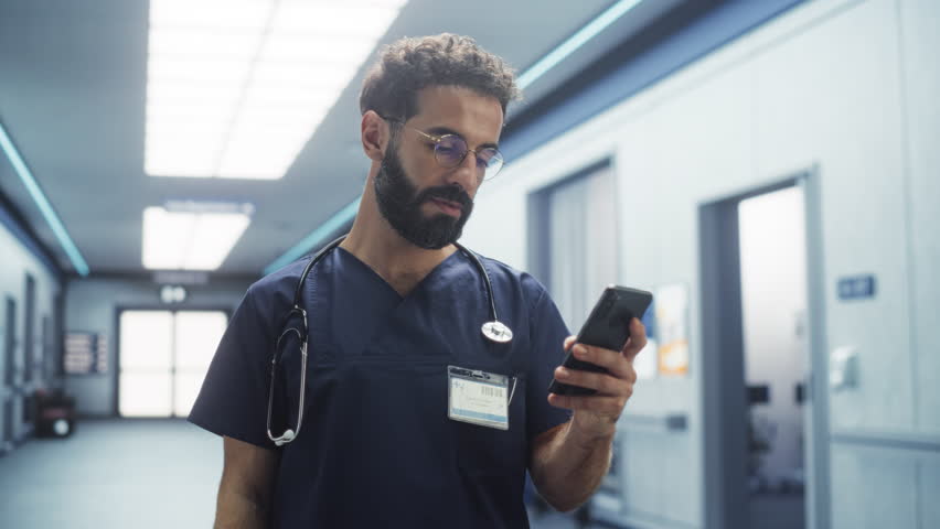Male Healthcare Nurse Using Smartphone to Check on his Messages and Emails During his Shift. Latin Young Clinic Professional Scrolling on Social Media During his Break in a Modern Hospital Corridor - Powered by Shutterstock - Get 15% off with code: PIKWIZARD15
