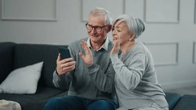 Senior couple having video call at home. Old gray haired man woman chatting talking on smartphone sitting on couch. Happy pensioners enjoying online conversation with family, friends smiling laughing.