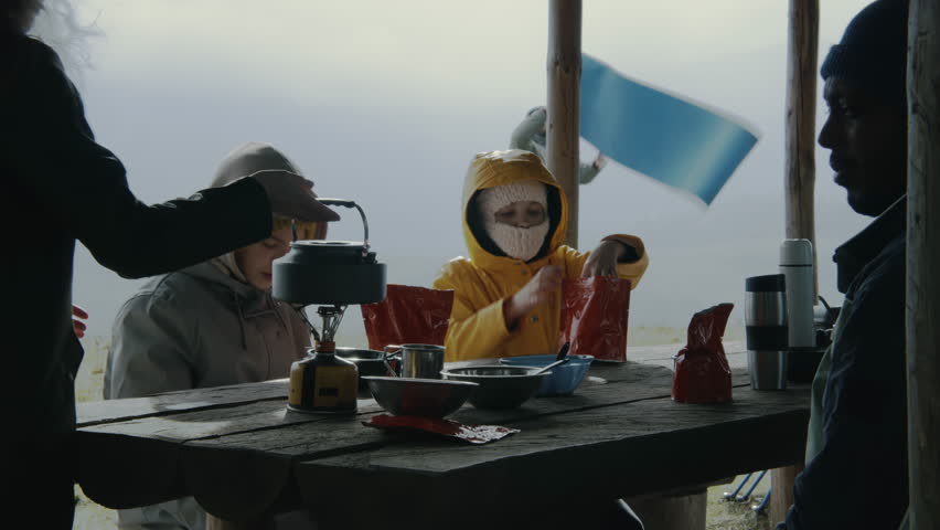 Group of diverse tourists rests in gazebo after long expedition. Young boy and girl wait for meal. Adult Caucasian woman takes kettle and cooks hiking food in pouch. Tourism and outdoor exploration.