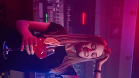 Pretty Young Happy Girl Smiles and Dances in Nightclub with Red Cocktail in her Hands. Background there is Bar Counter. Slow Motion, Vertical Video
