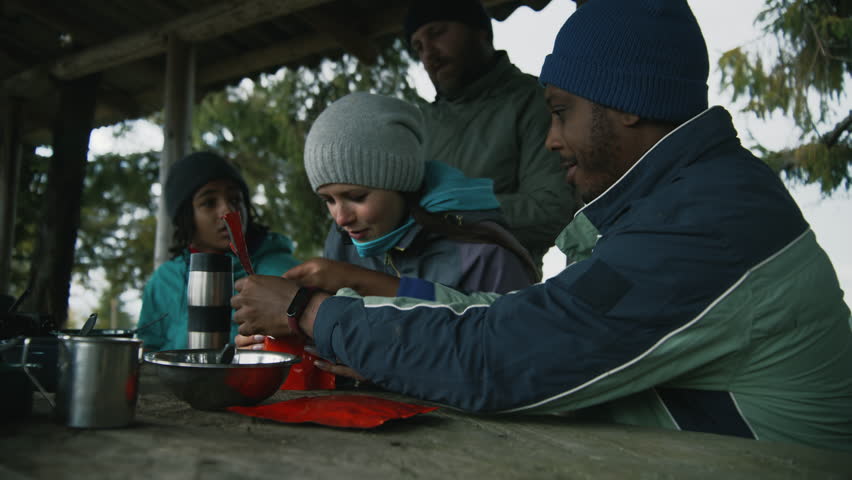 Adult African American man opens doy pack for his wife. Multiethnic tourist family eat hiking food together. Group of travelers or hikers rest in gazebo after expedition or trek in the mountains.