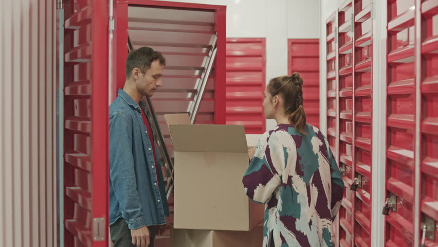 Young woman sealing box with adhesive tape while her husband putting it into locker room in self service storage