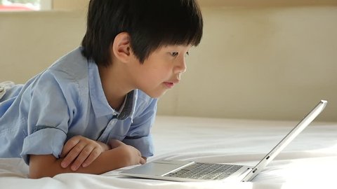 Cute Asian boy lying in bed and using laptop on white bed slow motion