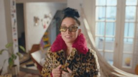 Young stylish girl in trendy fur coat with leopard print and sunglasses standing in sunlit room at home and looking at camera with happy smile. Zoom shot, video portrait