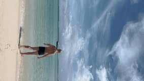 Shirtless man walking towards sea for swimming. Male tourist enjoying vacation during summer. Boats moored in ocean against sky, Vertical video