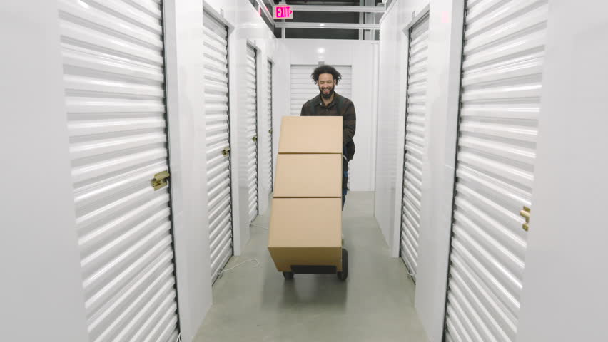 Happy bearded male African American pushing trolley cart with large carton boxes in front of him along white corridor in a climate-controlled storage. High quality 4k footage Royalty-Free Stock Footage #3404978571