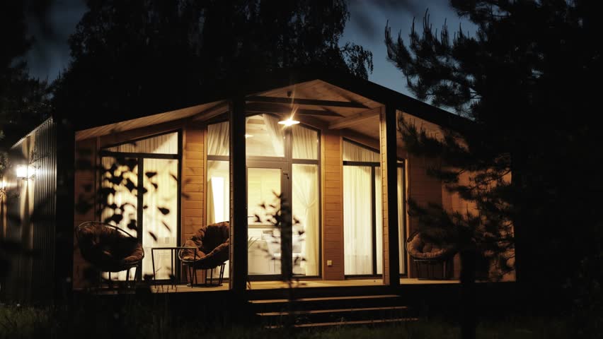 Night shot of wooden scandinavian house the light is on in the window, cozy terrace in summer with warm electric light. Forest cabin in the middle of a forest in silence, night sky, slow motion.