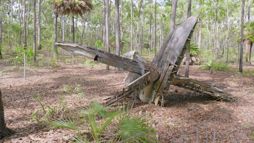 A wrecked Curtiss P-40e Warhawk plane crash in remote far north Australia.  There is a low fence surrounding the spread-out wreckage.  The surrounding thick bushland contrasts the rusted form.  In 4K.