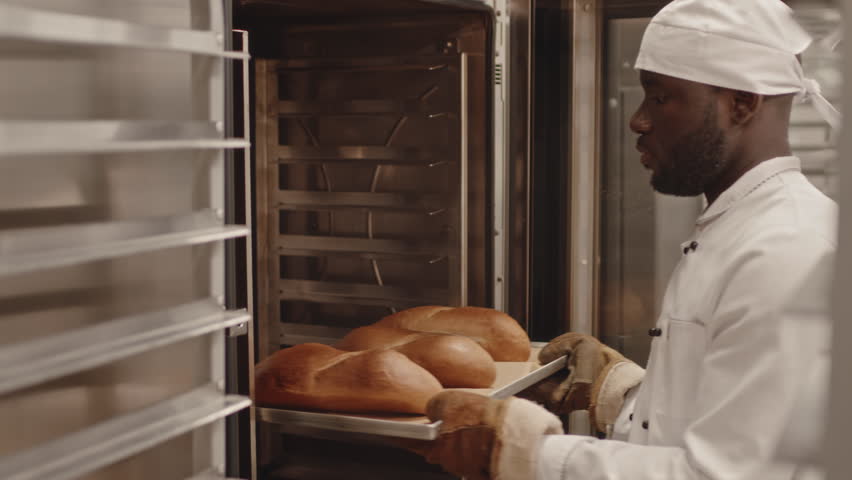 Medium shot of African American male bakery worker in white chef uniform taking baking tray with fresh loaves of bread out of oven and putting them onto tray rack Royalty-Free Stock Footage #3405132785
